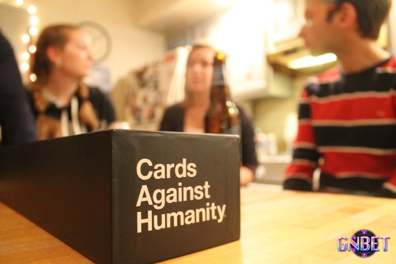 Nhóm bạn ngồi quanh bàn phòng khách buổi tối và bật cười khi thẻ đen vừa được đọc to trongboard game odd
